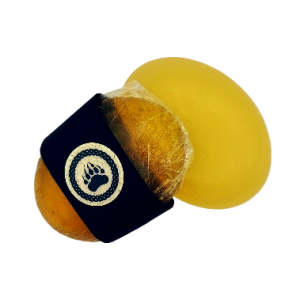Dos jabones artesanales ovalados de color ámbar dorado sobre fondo blanco. Uno está envuelto en plástico transparente y decorado con una banda negra que lleva el logo de Arkoni: una huella de oso dentro de un marco circular. El otro está sin envoltura, mostrando su textura suave y translúcida.